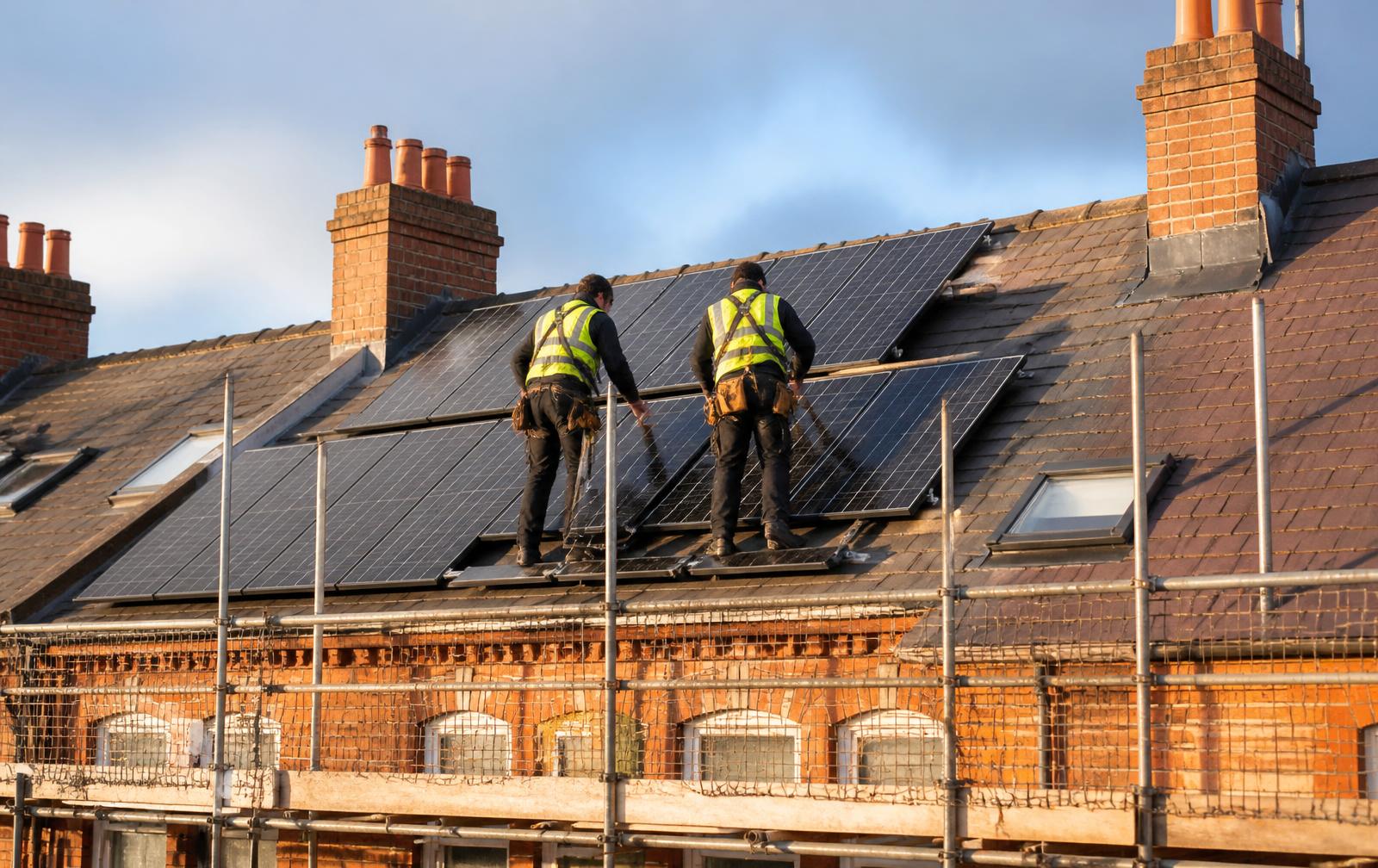 Two roofers in hi-vis and harnesses fitting solar panels to a slate roof on a brick terraced house